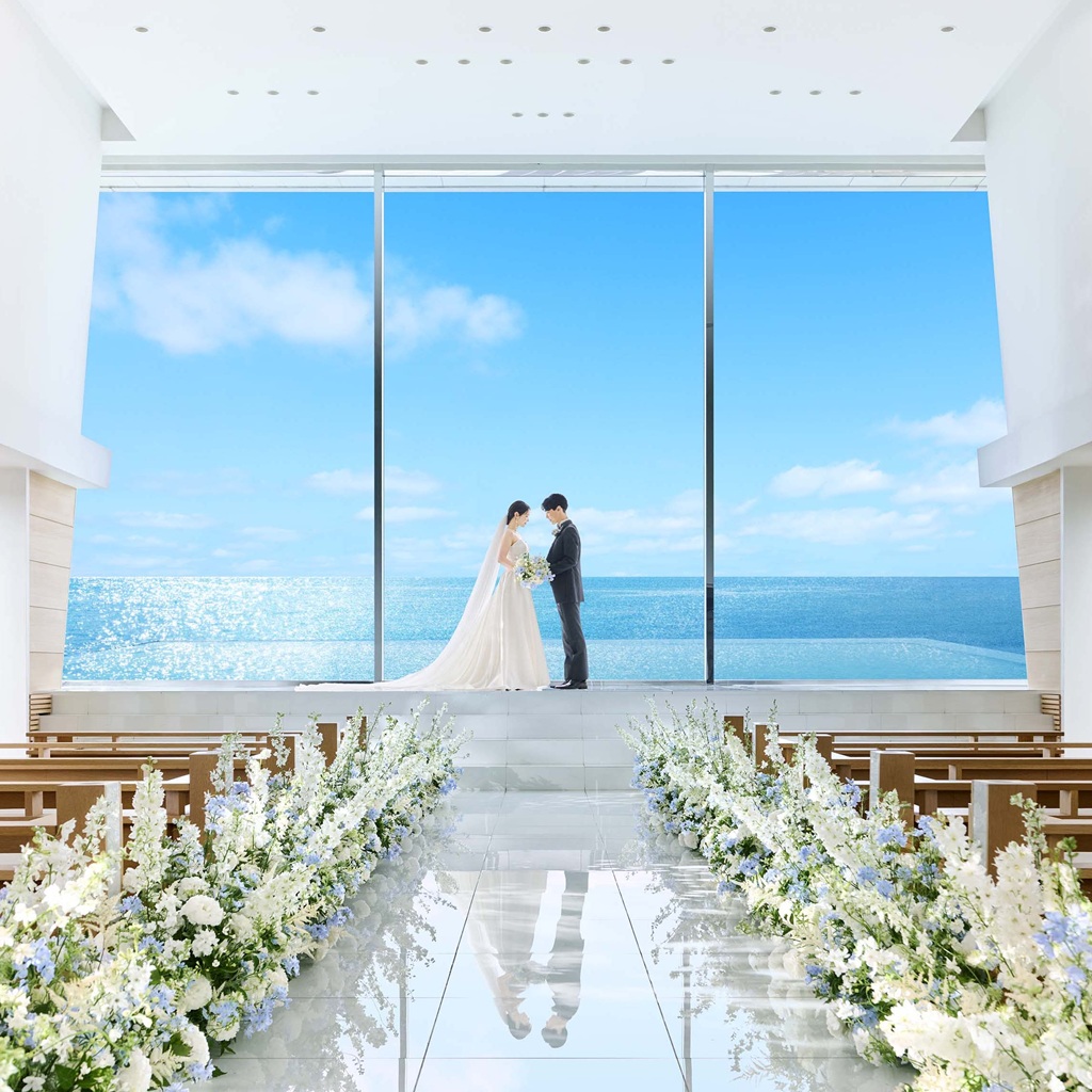 「海の青さが広がるガラス張りのチャペルで、祭壇に立つ新郎新婦が向かい合うウェディングシーン」